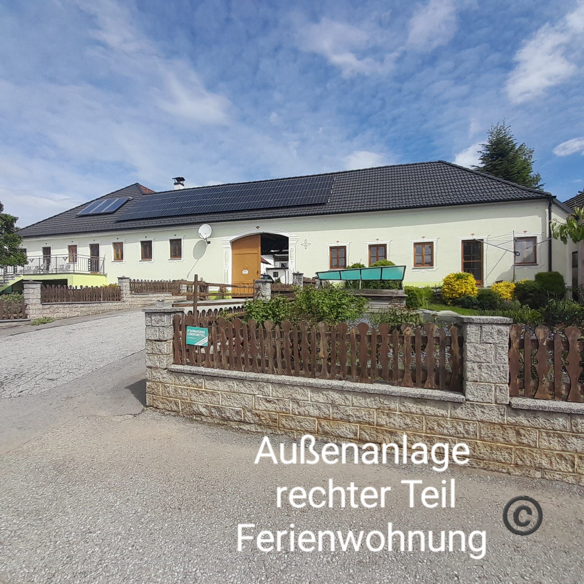 Ein großes Gebäude mit gelber Fassade und schwarzem Dach, umgeben von einem Garten und einem Holzzaun. Der Himmel ist blau mit einigen Wolken.