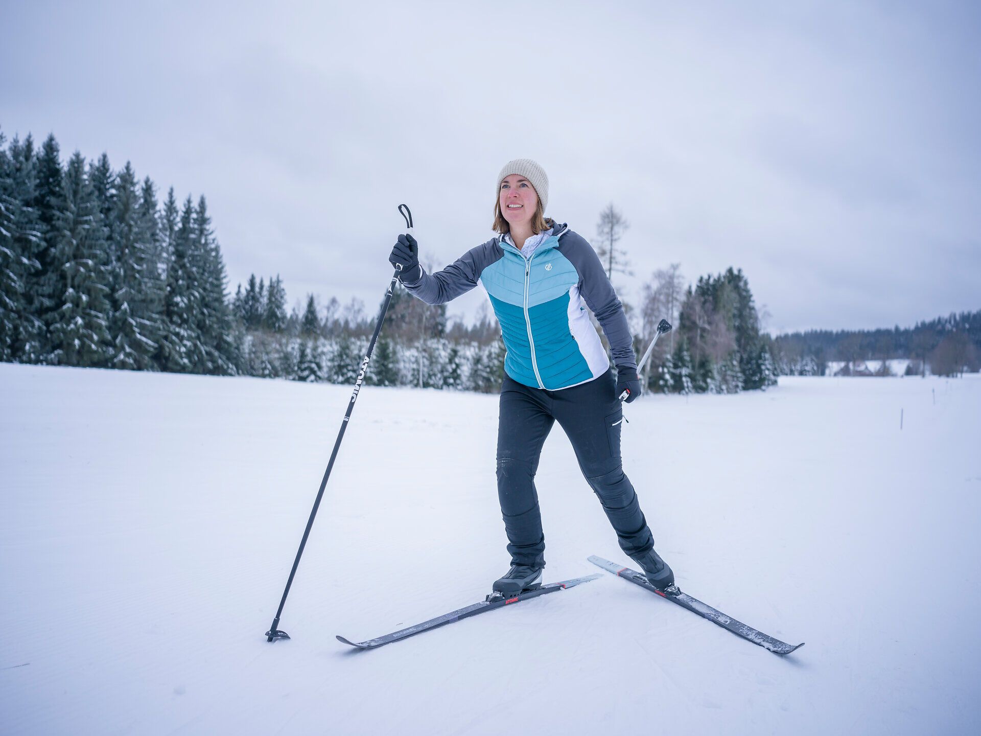 Eine Frau beim Langlaufen auf einer verschneiten Landschaft mit Bäumen im Hintergrund.