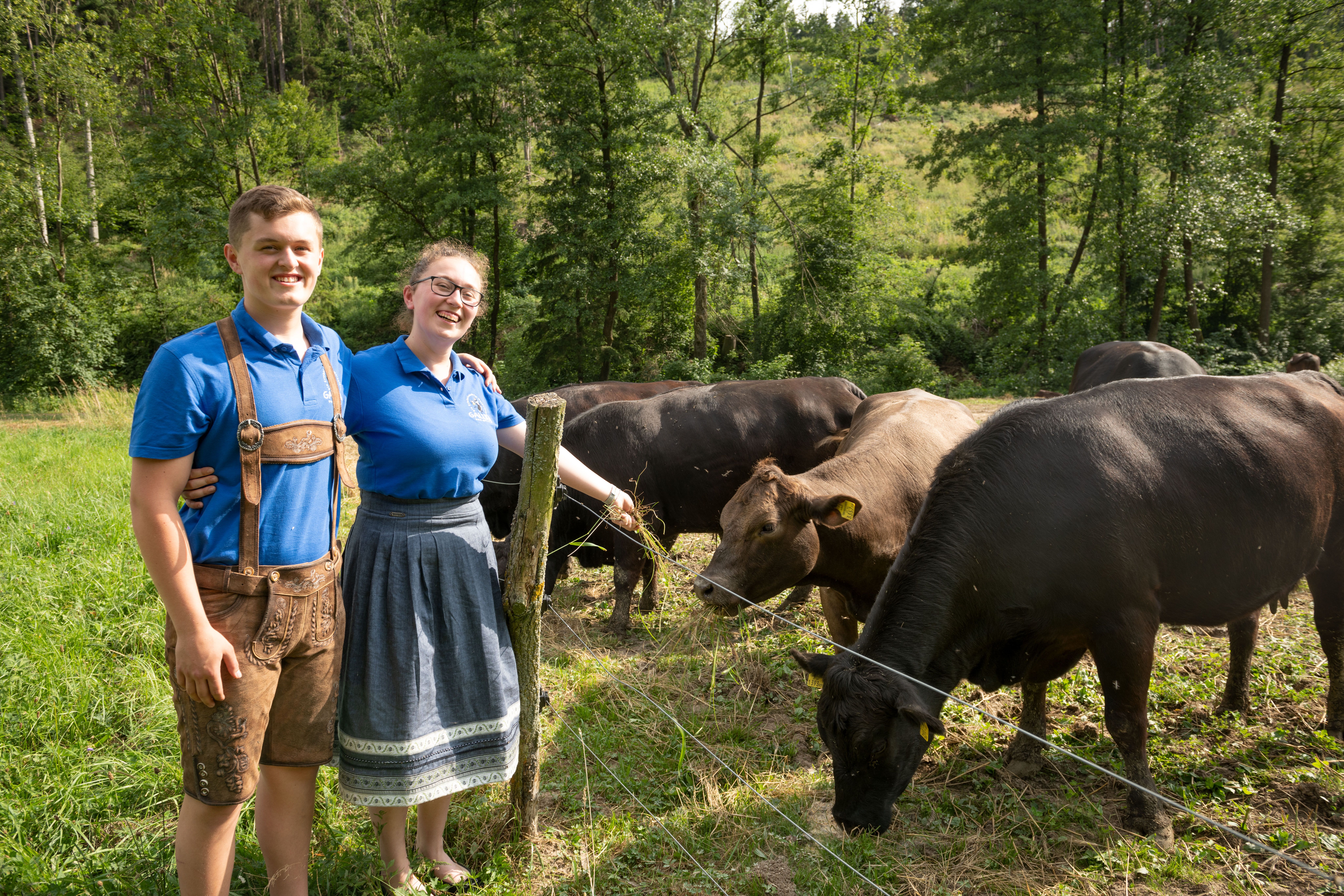 Zwei Personen in traditioneller Kleidung stehen neben einer Weide mit Kühen.