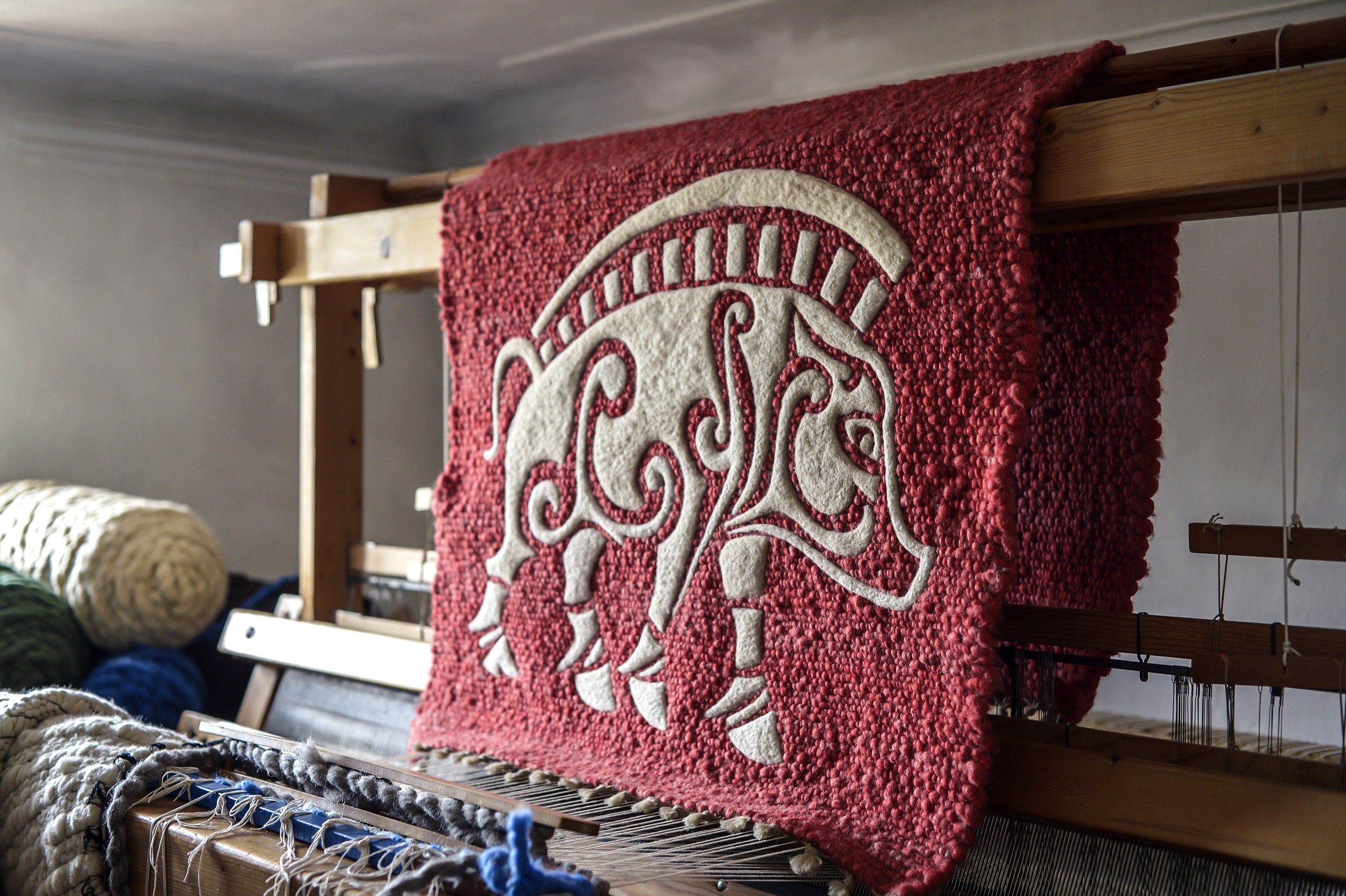 Ein roter Wandteppich mit einem weißen, stilisierten Tiermotiv auf einem Webstuhl.