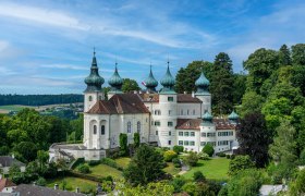 Ein weißes Schloss mit mehreren Zwiebeltürmen, umgeben von grüner Landschaft und Bäumen, unter einem blauen Himmel.