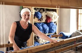 Frau am Webstuhl mit Wollknäueln im Hintergrund.