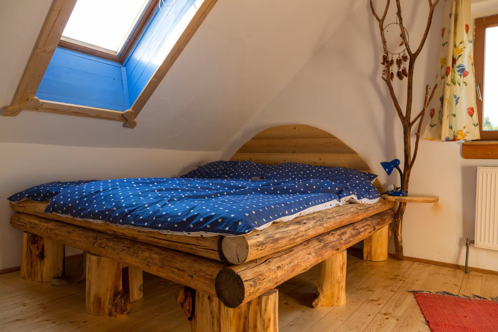 Rustikales Schlafzimmer mit Holzbett, blau gepunkteter Bettwäsche, Dachfenster und dekorativem Ast als Kleiderständer.