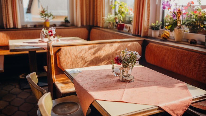 Gemütliche Stube mit Holzmöbeln, Tischdecken und Blumen auf den Tischen, umgeben von Fenstern mit Pflanzen.