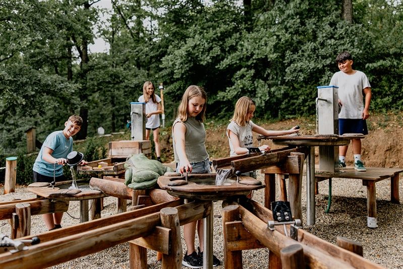 Kinder spielen an einem Wasserspielplatz im Freien, umgeben von Bäumen.
