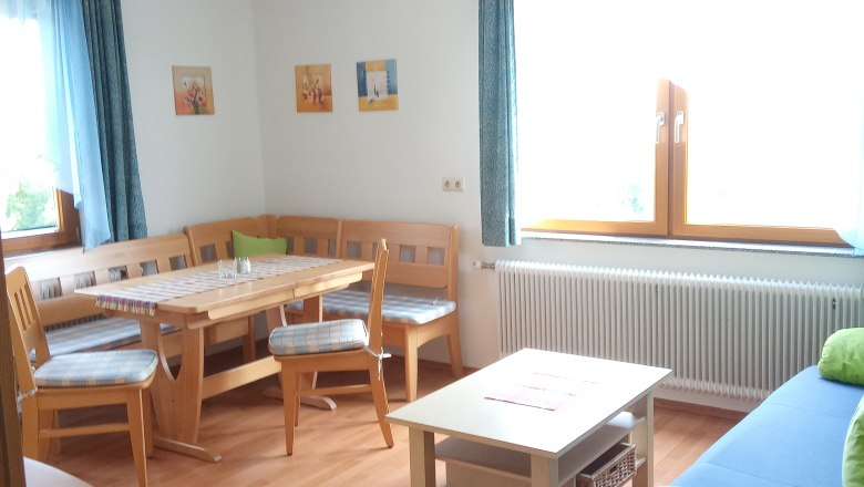 Helles Wohnzimmer mit Holzmöbeln, Eckbank, Tisch, Sofa und großen Fenstern mit blauen Vorhängen.