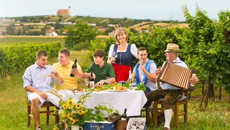 Familie sitzt an einem Tisch im Weinberg, trinkt Wein und genießt die Landschaft.