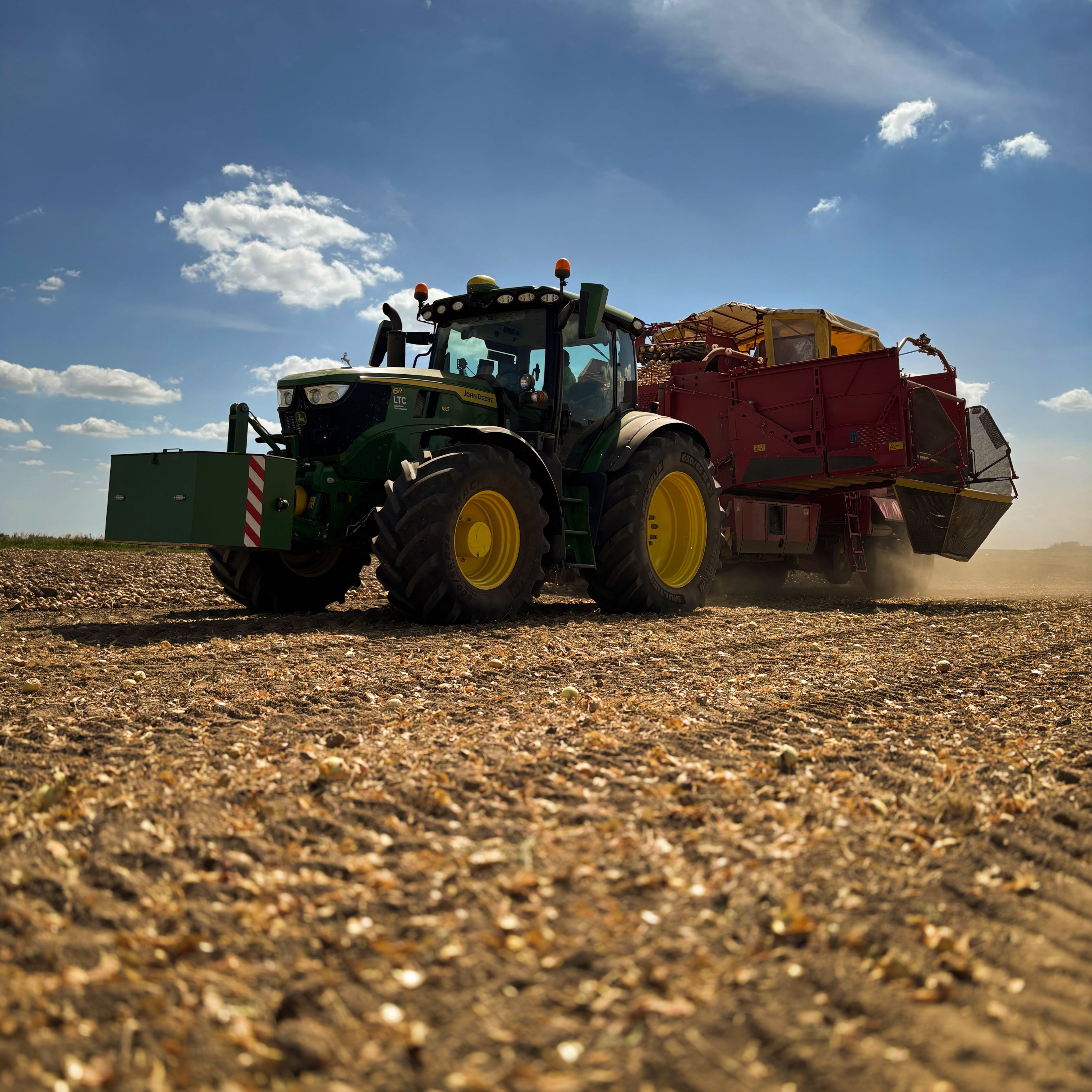 Ein Traktor mit Anhänger fährt über ein abgeerntetes Feld unter einem blauen Himmel mit einigen Wolken.
