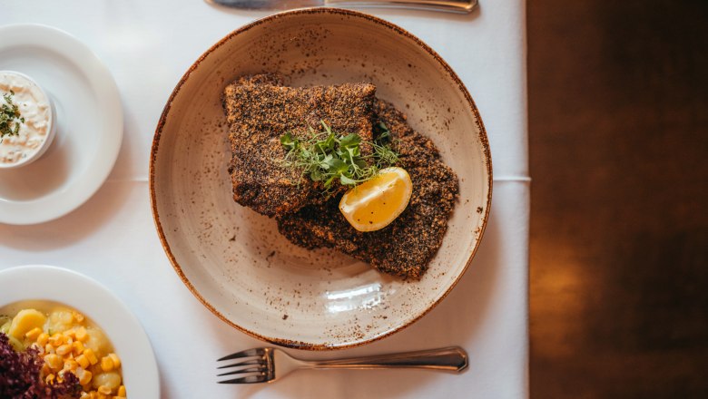 Graumohn-paniertes Karpfenfilet mit Zitronenscheibe und Kr&auml;utern auf Teller, daneben Beilagen.