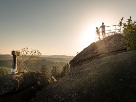 Die goldene Abendsonne taucht die majest&auml;tischen Felsen in ein warmes Licht, w&auml;hrend zwei Wanderer die atemberaubende Aussicht genie&szlig;en. Umgeben von der unber&uuml;hrten Natur und dem sanften Rauschen der B&auml;ume, l&auml;dt dieser Ort dazu ein, die Seele baumeln zu lassen und die Sch&ouml;nheit der Berge zu erleben.