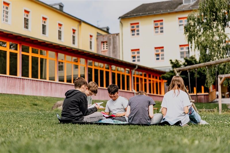 Gruppe von Kindern sitzt auf einer Wiese vor einem gelben Gebäude.