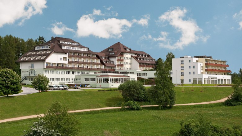 Großes Gebäude mit mehreren Stockwerken und Balkonen, umgeben von grüner Landschaft und Bäumen, unter blauem Himmel.
