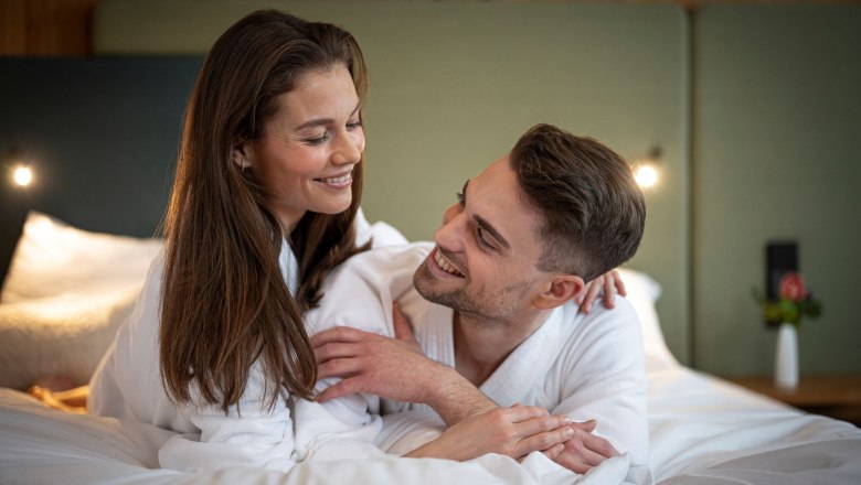 Ein lächelndes Paar in weißen Bademänteln liegt auf einem Bett und schaut sich an.
