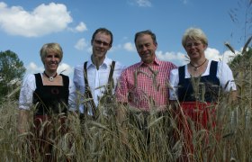 Vier Personen in traditioneller Kleidung stehen in einem Getreidefeld unter blauem Himmel.