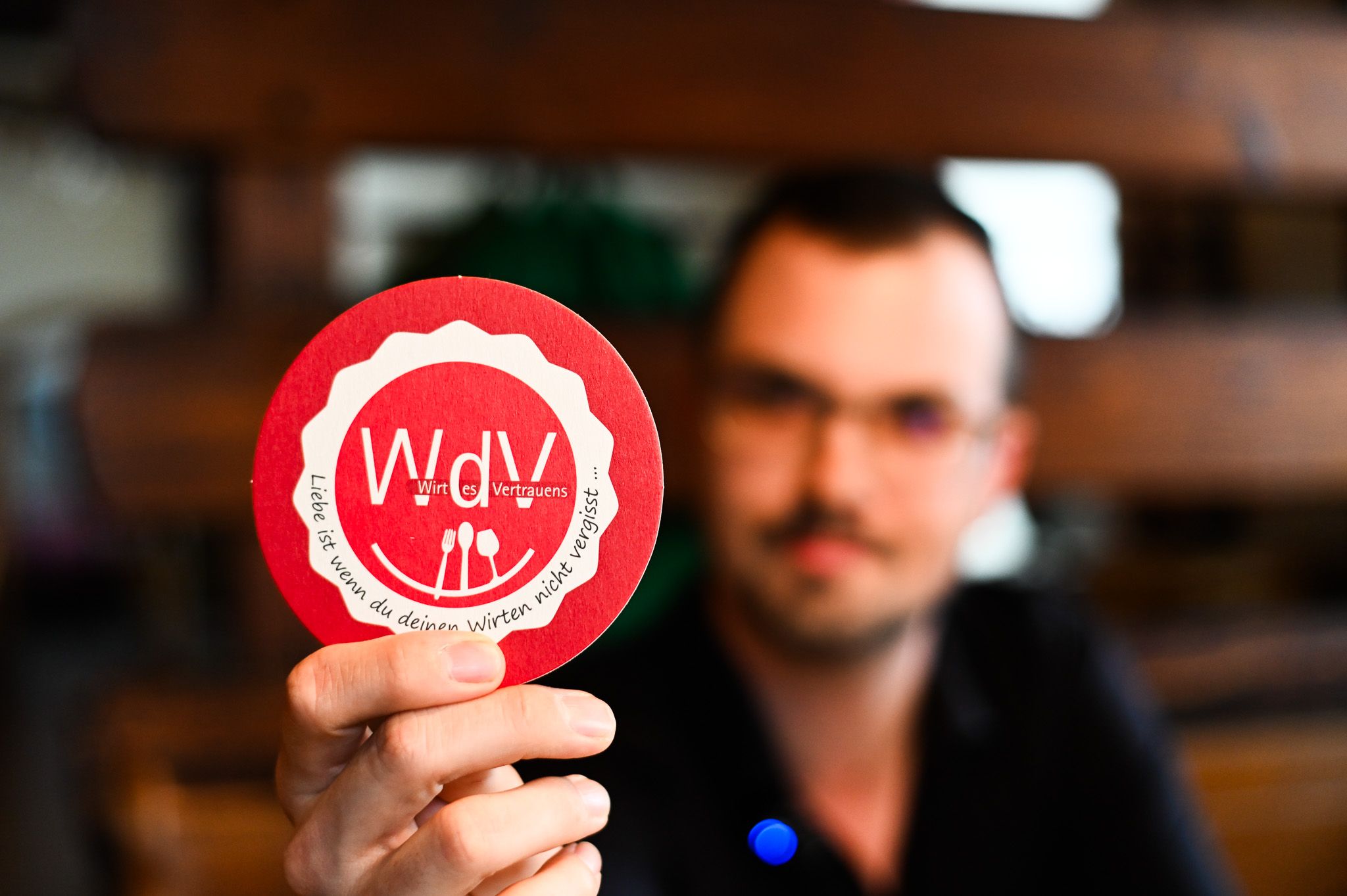 Ein Mann hält einen Bierdeckel mit der Aufschrift 'WdV - Wirt des Vertrauens' in die Kamera.