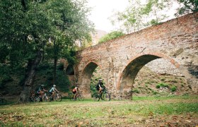 Gruppe von Radfahrern unter einer alten Backsteinbrücke im Grünen.