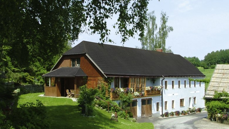 Ein traditionelles Bauernhaus mit Blumen auf dem Balkon, umgeben von gr&uuml;ner Landschaft und B&auml;umen.