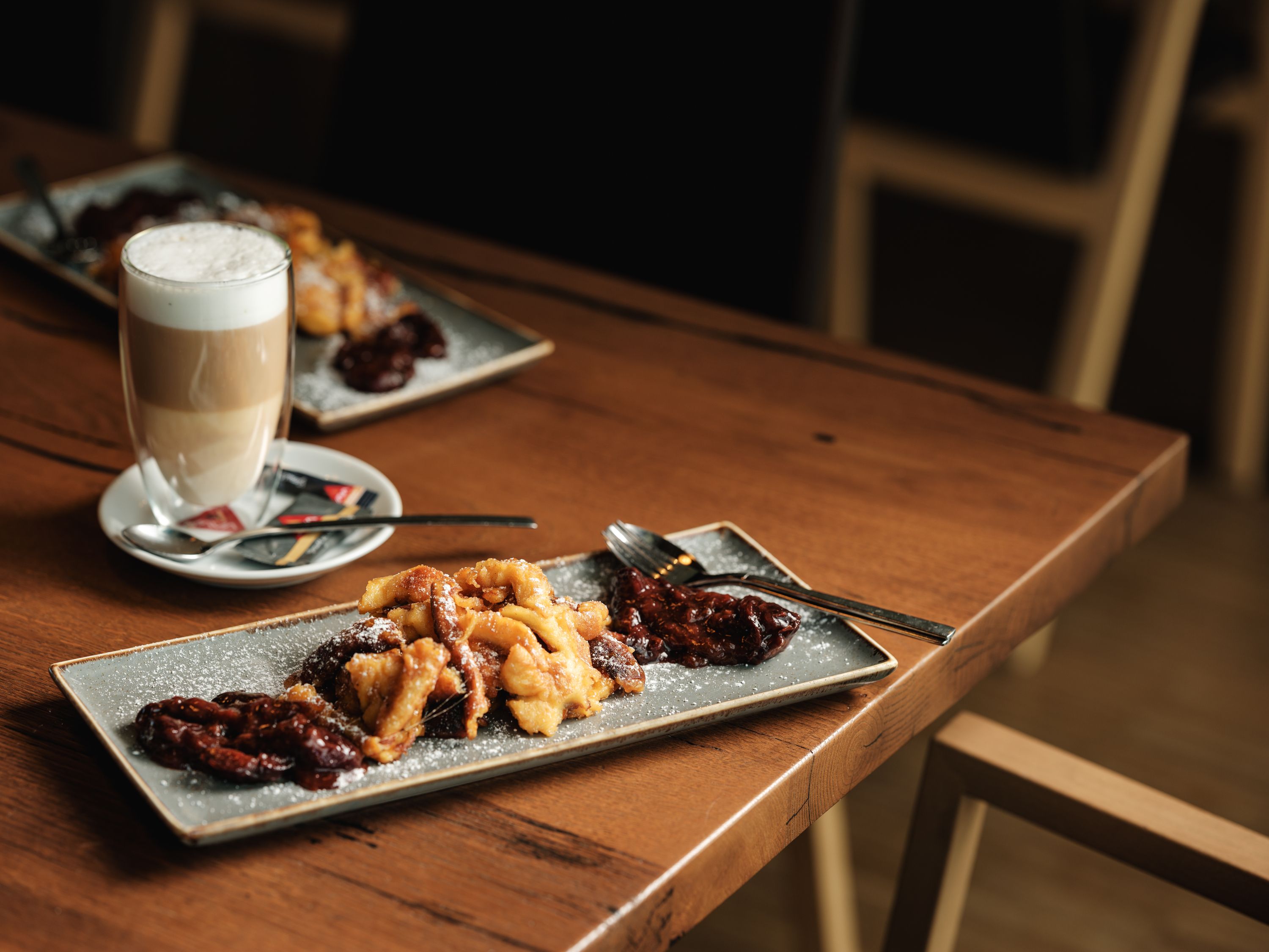 Ein Teller mit Kaiserschmarrn und Pflaumenkompott auf einem Holztisch, daneben ein Glas Latte Macchiato.