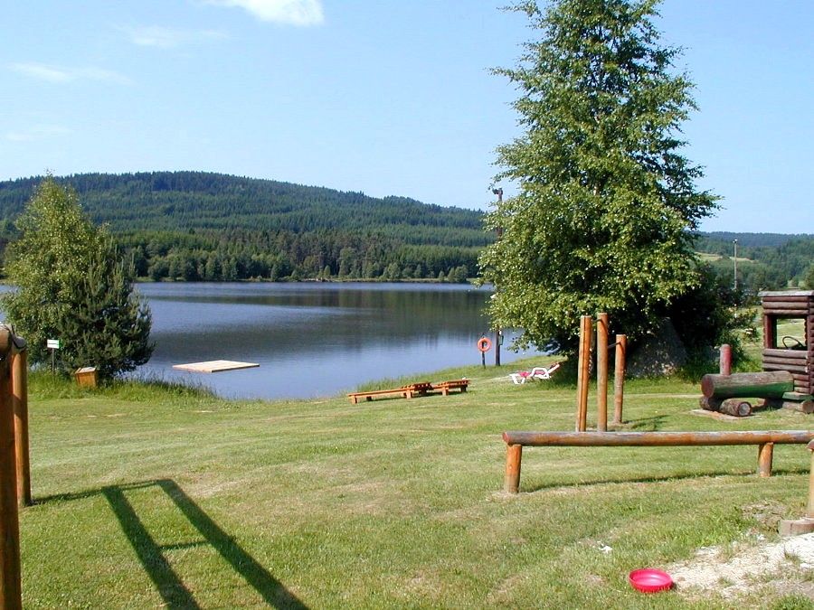 Ein ruhiger See mit Steg, umgeben von grünen Hügeln und Bäumen, mit einem Spielplatz im Vordergrund.