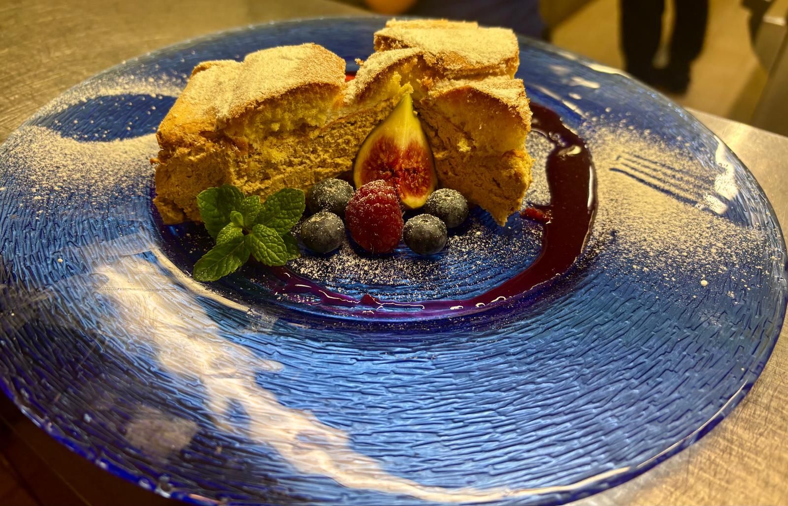 Ein Dessert auf einem blauen Teller, dekoriert mit Beeren, Feige und Minzblättern, bestäubt mit Puderzucker.