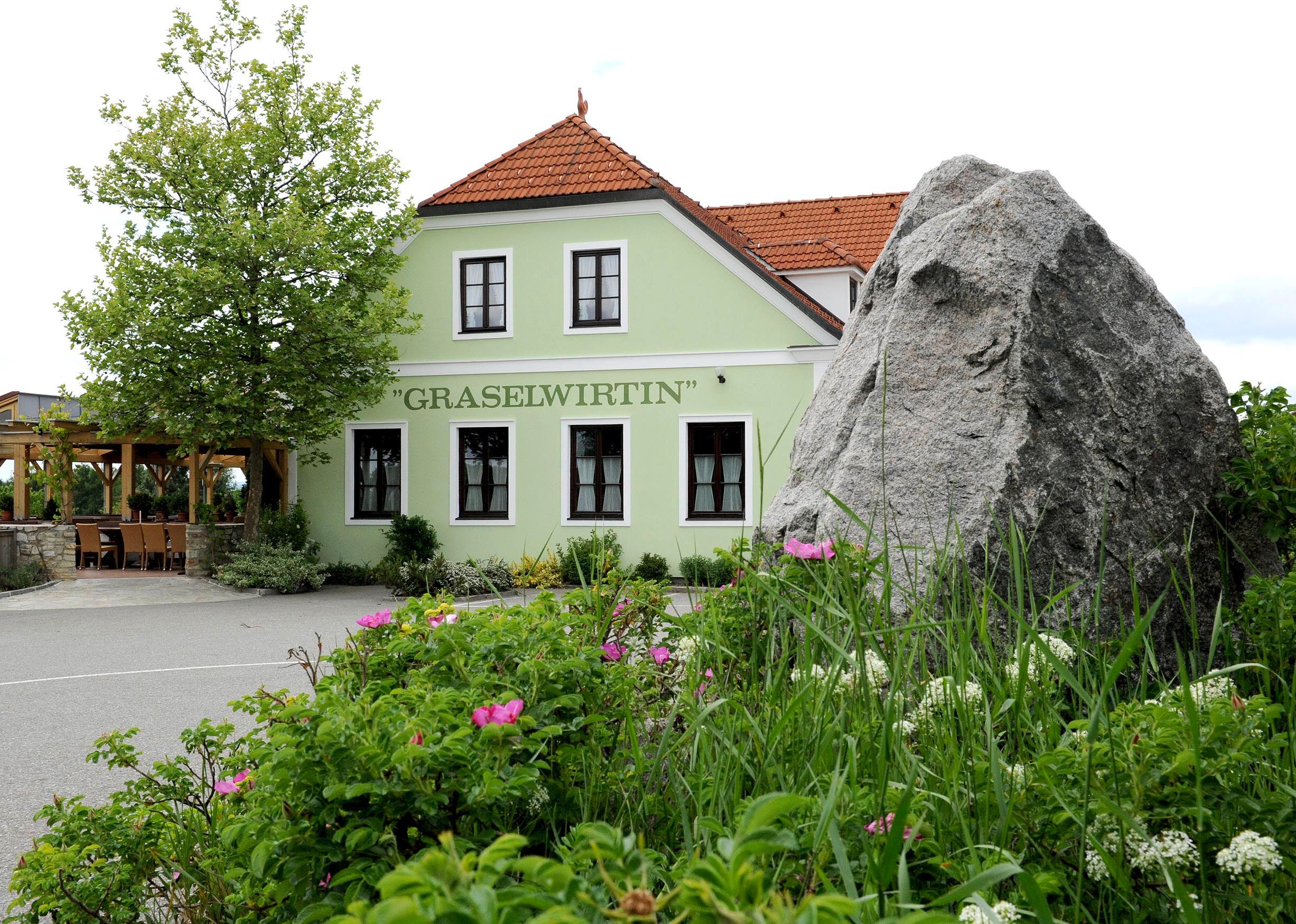 Grünes Gebäude mit der Aufschrift 'Graselwirtin', umgeben von Pflanzen und einem großen Felsen im Vordergrund.