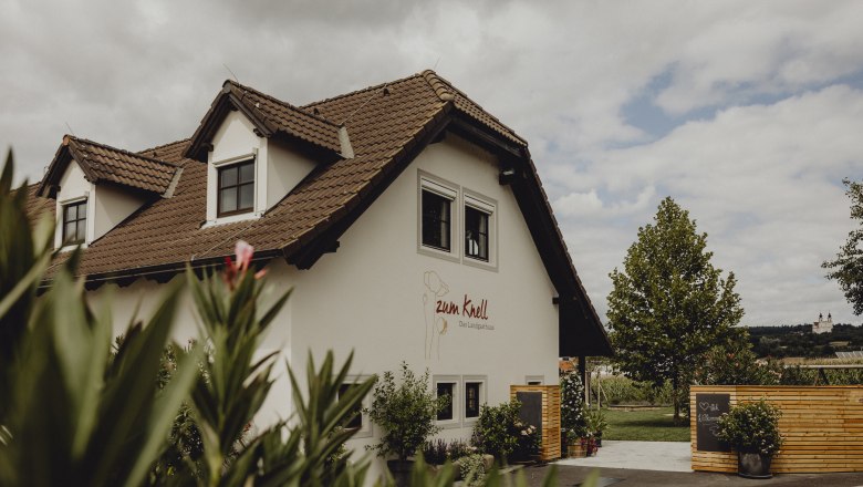 Ein Landgasthaus mit braunem Ziegeldach und weißer Fassade, umgeben von Pflanzen und einem Holzzaun.