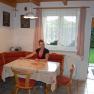 A woman sits at a dining table in a cozy kitchen with a corner bench and red cushions.