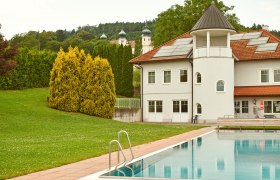 Außenansicht einer Freizeitanlage mit Pool und Gebäude, im Hintergrund Bäume und Kirchtürme.