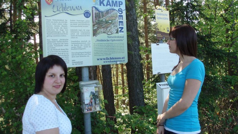 Zwei Frauen stehen vor einem Schild mit der Aufschrift 'Naturdenkmal Heidnische Opferstätte' im Wald.