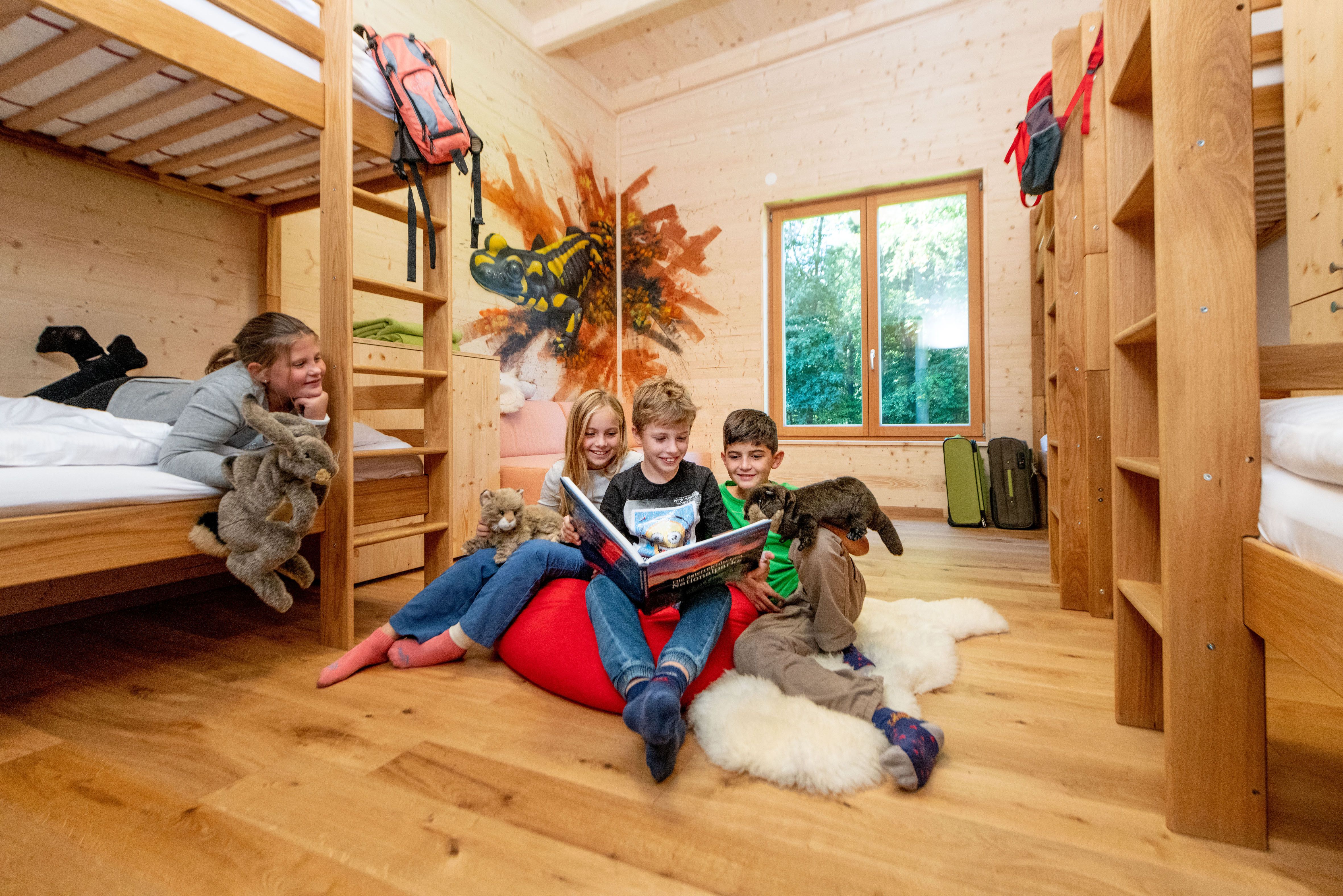 Kinder in einem Holzzimmer mit Etagenbetten, lesen ein Buch und halten Plüschtiere.