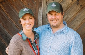 Zwei Personen l&auml;cheln in die Kamera, tragen gr&uuml;ne Kappen mit einem Logo und stehen vor einer Holzwand.