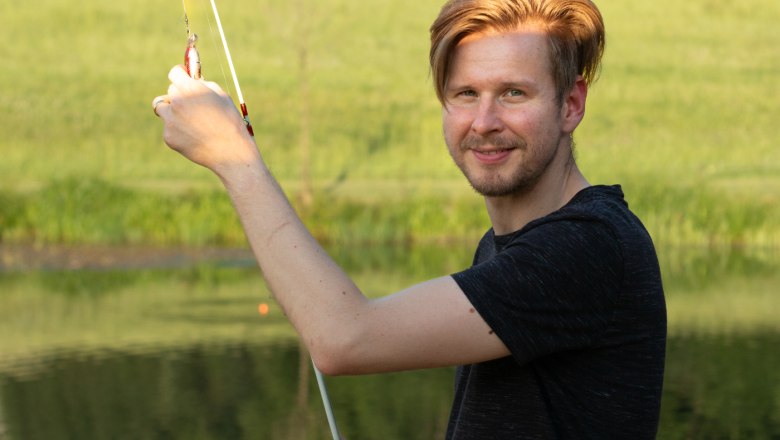 Mann mit Angelrute am Teich vor grüner Landschaft.