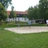 Menschen spielen Volleyball auf einem Sandplatz im Freien, umgeben von Bäumen und einem Gebäude im Hintergrund.