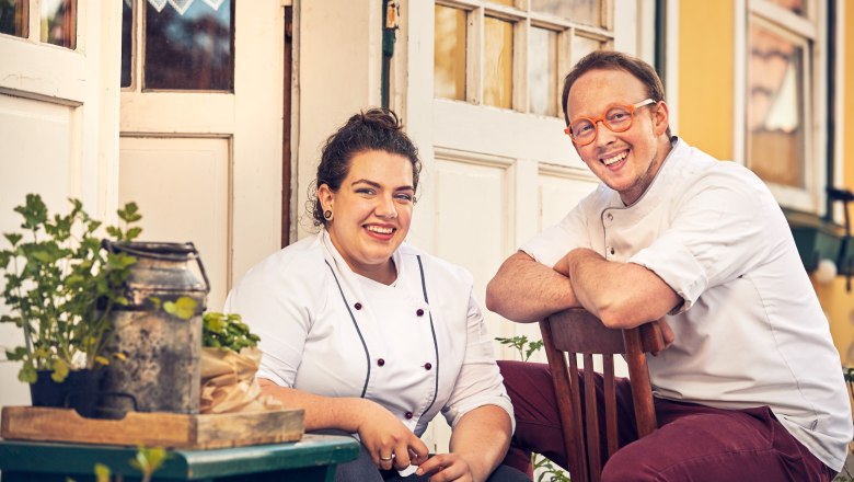 Two people in chef's clothing sit smiling in front of a building.