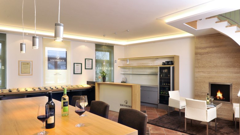 Innenansicht eines modernen Weinverkostungsraums mit Holztisch, Weingl&auml;sern, Kamin und Regalen.