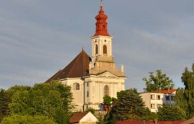 Wallfahrtskirche auf einem Hügel mit roten Dächern und Bäumen im Vordergrund.
